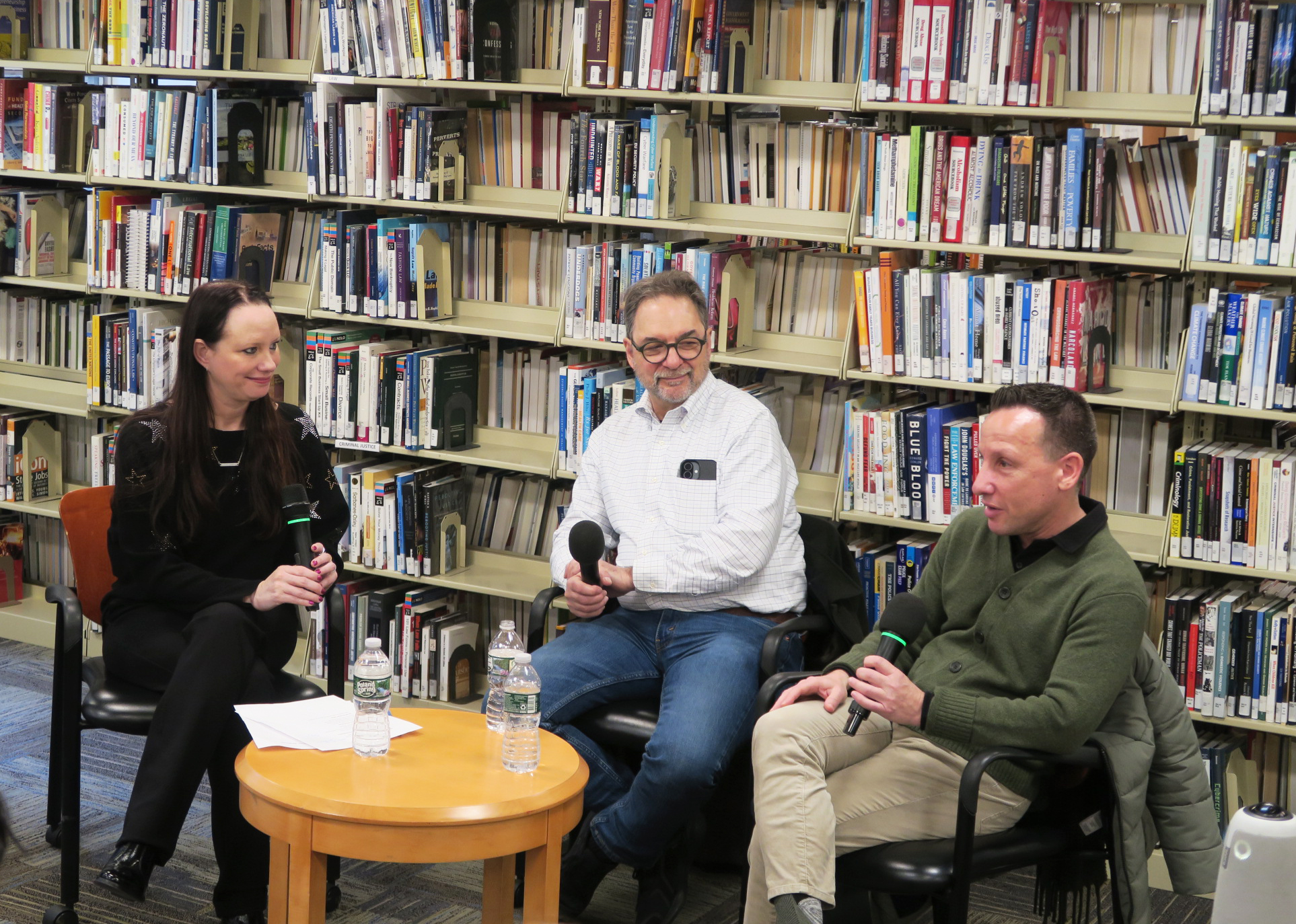 a woman and two men sit at a table in front of a shelf of books