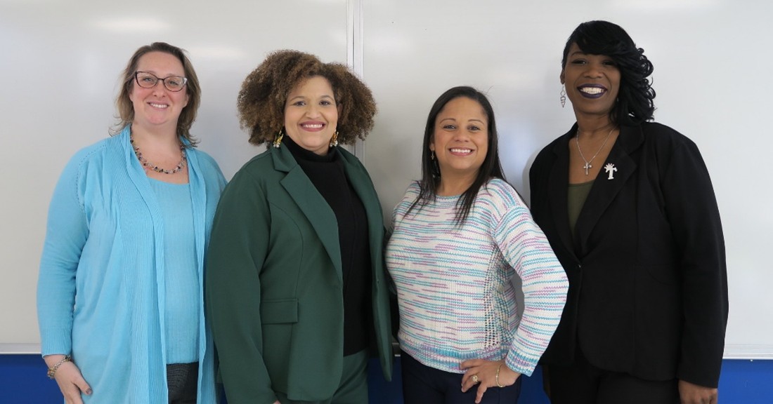 Four diverse women stand smiling in front of a whiteboard.