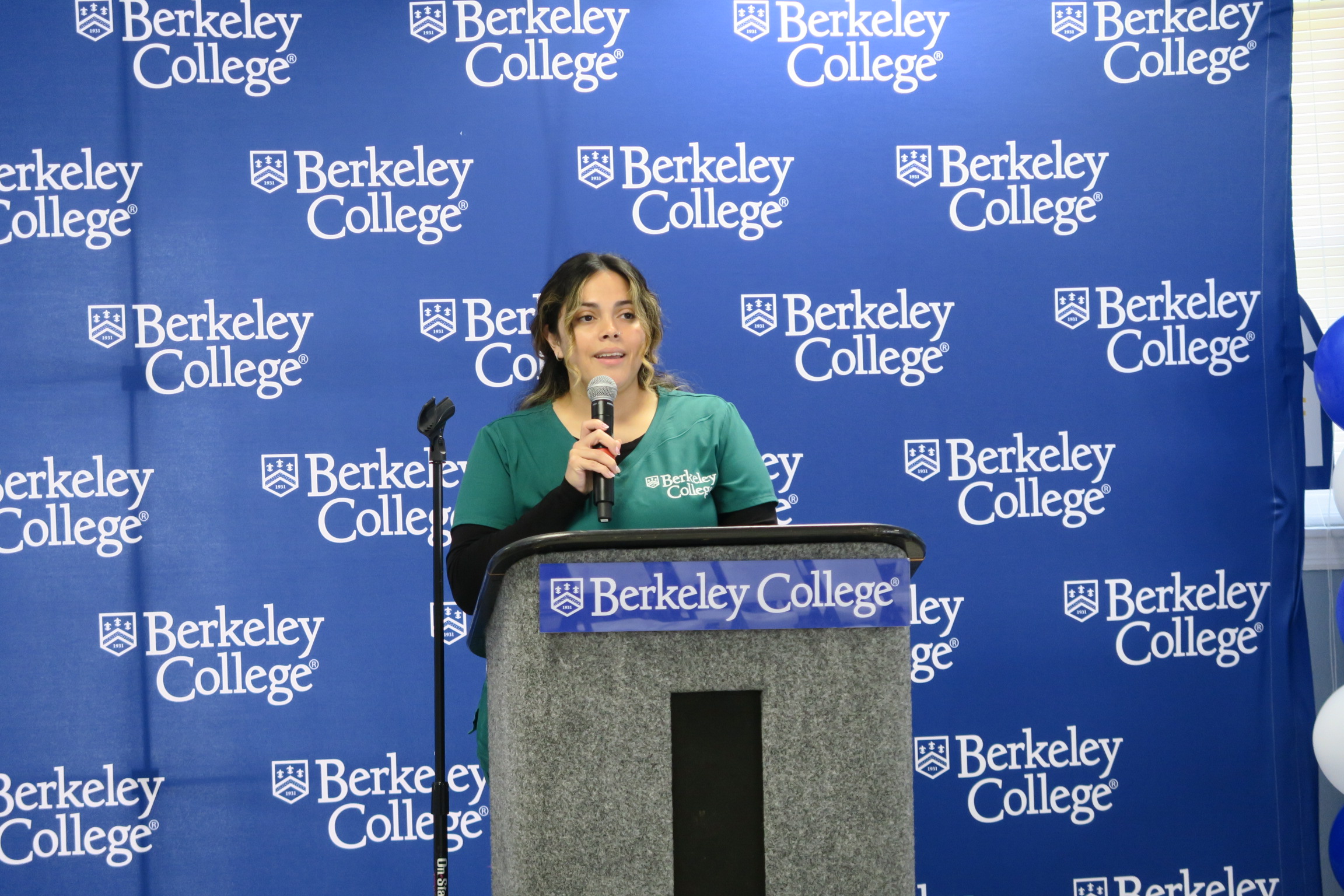 A female nursing student in scrubs gives a speech at a podium holding a microphone.