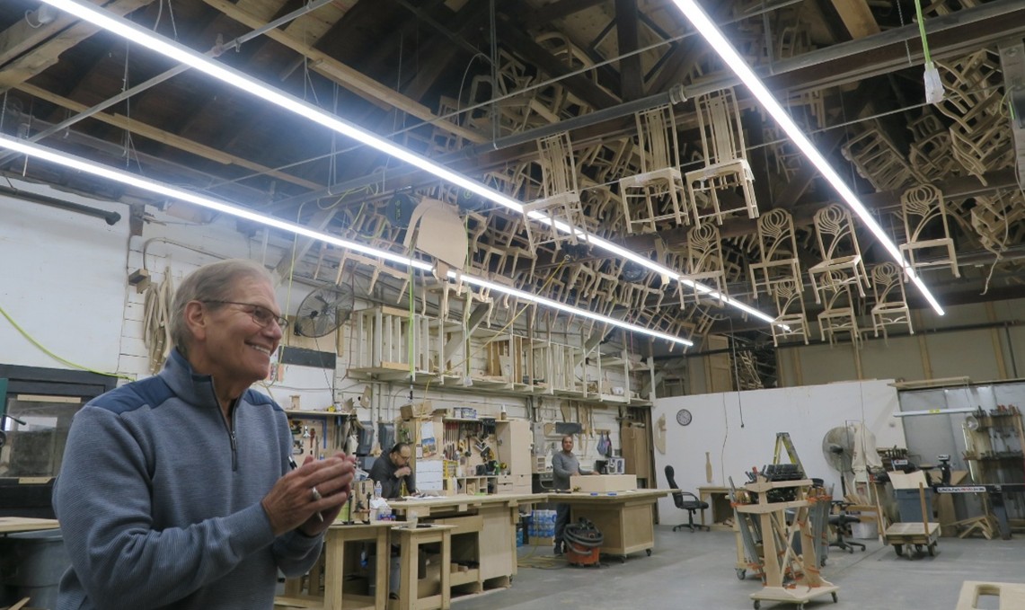 A man a blue sweater with folded hands smiles in an open warehouse with chairs hanging from the ceiling.